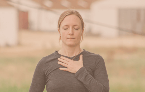 Woman with eyes closed and one hand on her chest, practicing a calming breath technique outdoors, symbolizing nervous system regulation and inner safety.