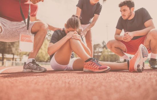 Female athlete sitting on a track clutching her knee in pain, surrounded by teammates and a coach, symbolizing sports injury, overtraining, and the consequences of ignoring rest.
